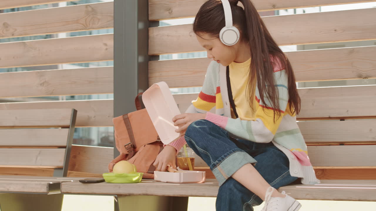 Asian Schoolgirl in Headphones Eating Sandwich Outdoors