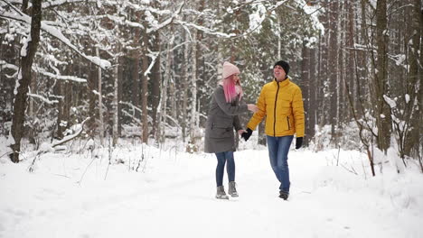 a man in a yellow jacket and a girl in a hat and scarf walk through the winter forest during a snowfall laughing and smiling at each other at christmas in slow motion