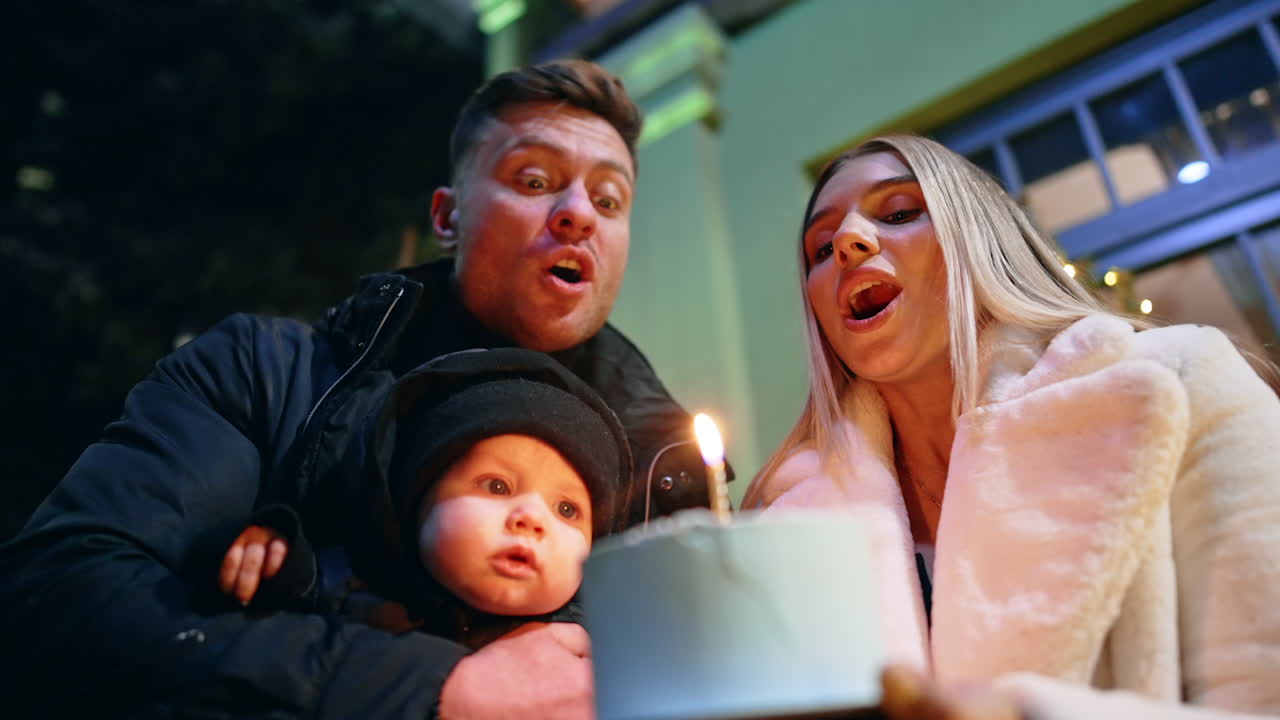 Happy Caucasian family making a wish. Dad holds an infant son and woman holds the cake with candle. Parents blow the candle together. Low angle view.