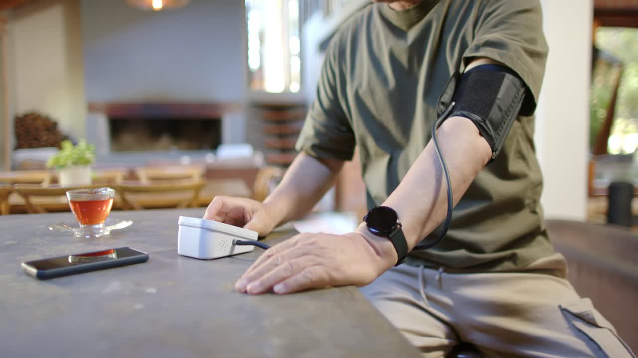 Anziano uomo asiatico che preme il monitor della pressione sanguigna, stringendo il bracciale sul braccio sinistro al tavolo da pranzo, controllando la sua salute