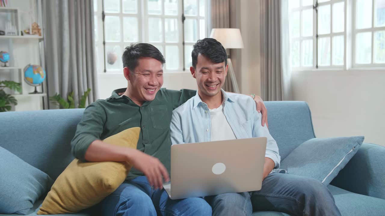 Portrait Of Asian Gentle Gay Couple Using Laptop Computer, While Sitting On A Couch In Cozy Stylish Apartment. Adult Boyfriends Online Shopping On Internet, Watching Funny Videos On Streaming Service