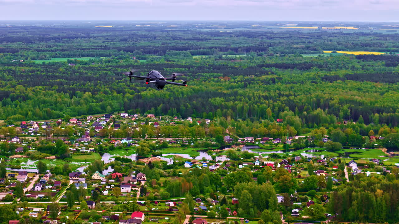Black civilian drone flying above town surrounded by forest and farmland during spring