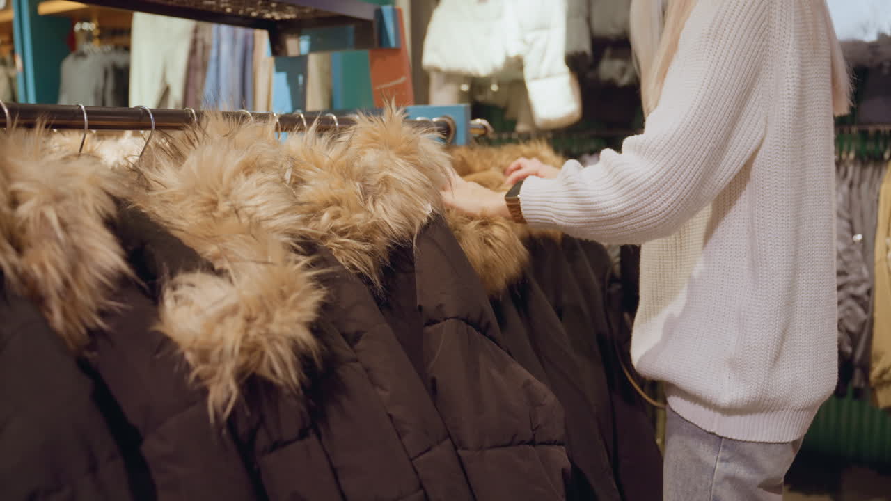 Trend setter browsing coat rack feeling fur trim assessing style and quality under warm boutique lighting with shoes displayed on top amid organized winter apparel racks evoking shopping ambience