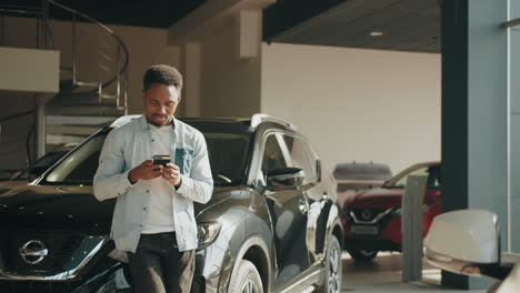 hombre mirando su teléfono mientras está de pie junto a un coche en un concesionario