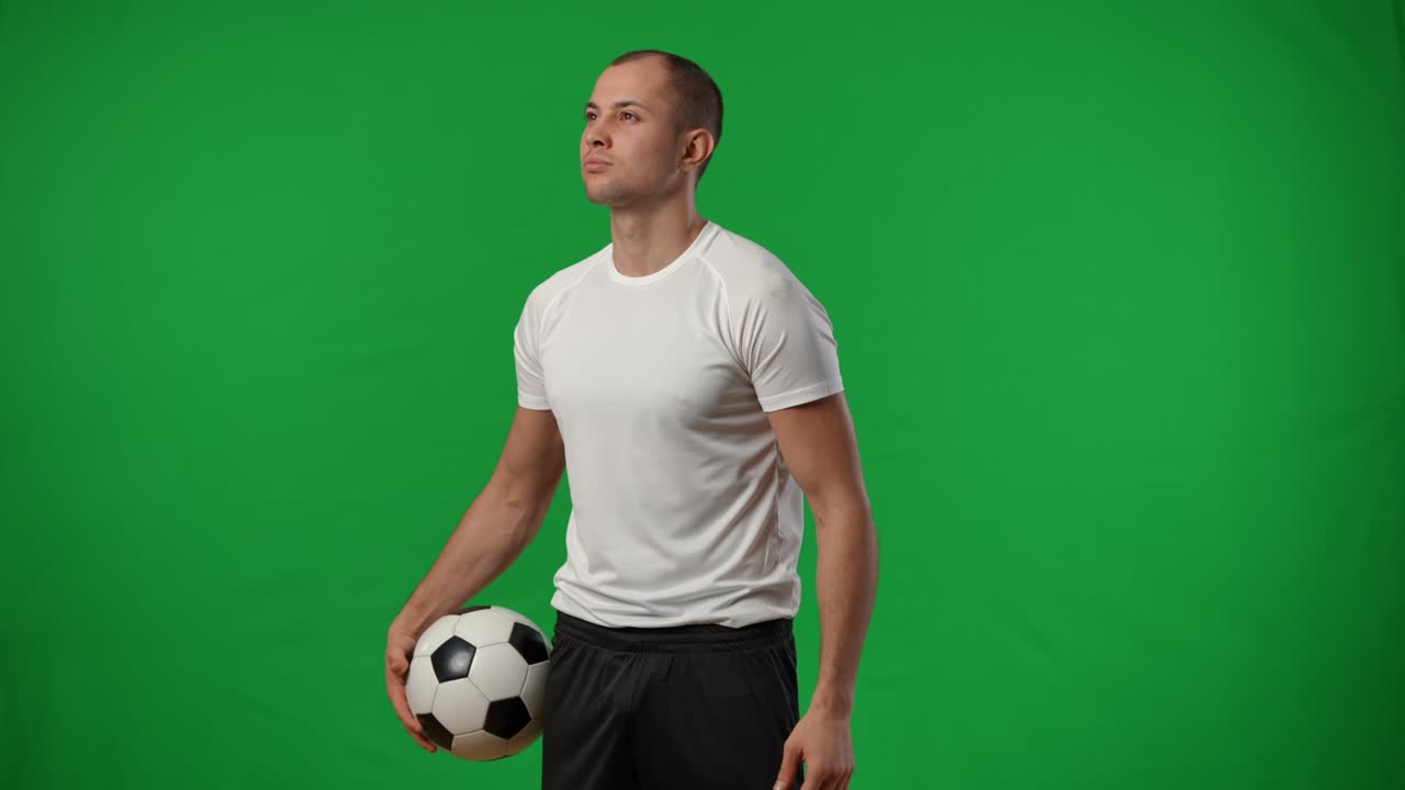 joven futbolista caucásico confiado y positivo con una pelota de fútbol de pie en el fondo de chromakey mirando a su alrededor. retrato de un deportista exitoso satisfecho y sonriente en la pantalla verde.