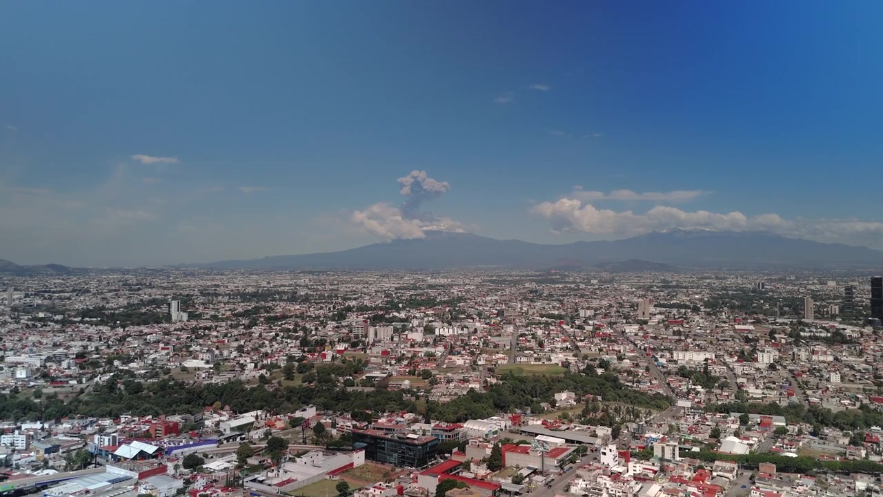 Vista panoramica della città di Puebla con il maestoso vulcano Popocatépetl che si erge in lontananza e rilascia fumo vulcanico. Un paesaggio messicano visivamente accattivante che combina architettura urbana e elementi naturali.