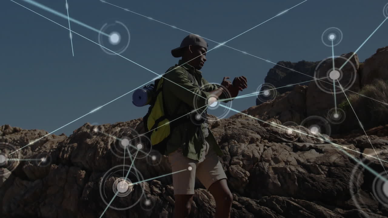 Male hiker standing on rocky outcrop, checking smartwatch showing digital network nodes for tech