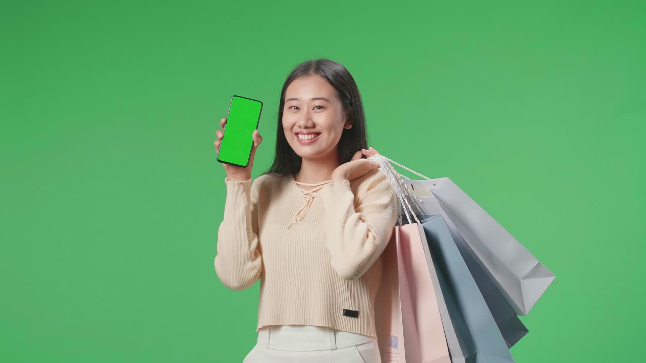 An Asian Shopping Woman Holding Green Screen Mobile Phone And Shopping Bags Smile To Camera While Standing In Front Of Green Screen