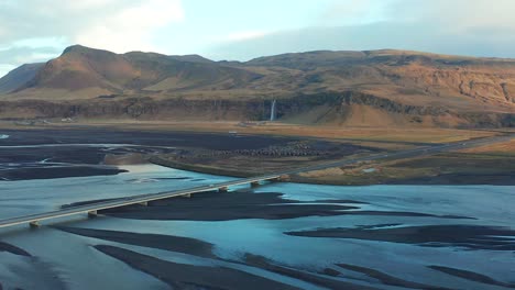 Aerial-view-of-a-river-estuary-being-cross-by-a-bridge,-Iceland-panoramic-view-of-Seljalands-with-mountainous-Seljalandsfoss-cascade