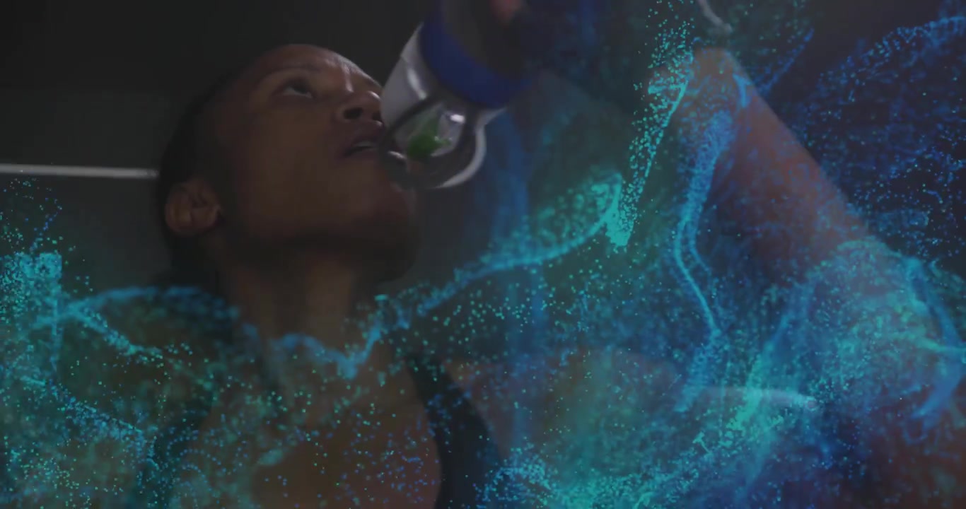 Drinking woman lifting water bottle at gym, wearing dark tank top and wrist glove, teal particles