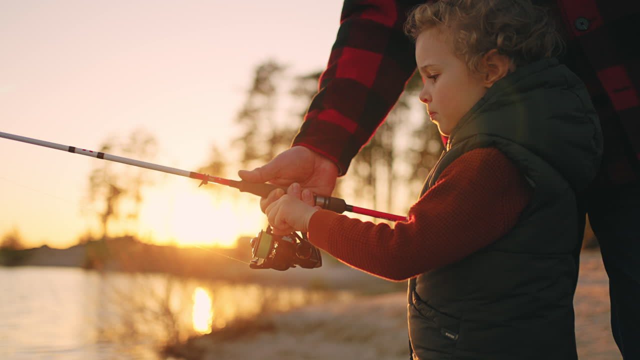 fishing on shore of river or lake in sunset little boy is catching fish by rod father or granddad is helping