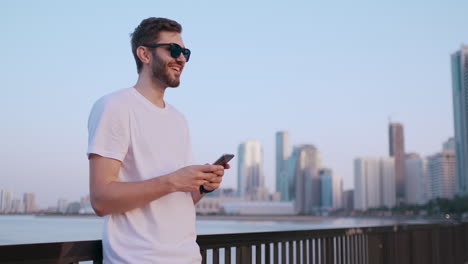 Attraktiver-Und-Gutaussehender-Junger-Mann-Mit-Schönem-Lächeln-Und-Hipster-Look-Der-Millennial-Generation-Z-Scrollt-Durch-Den-Social-Media-Feed-Auf-Dem-Smartphone,-Kichert-Und-Lächelt-Bei-Den-Nachrichten-In-Den-Anwendungen.