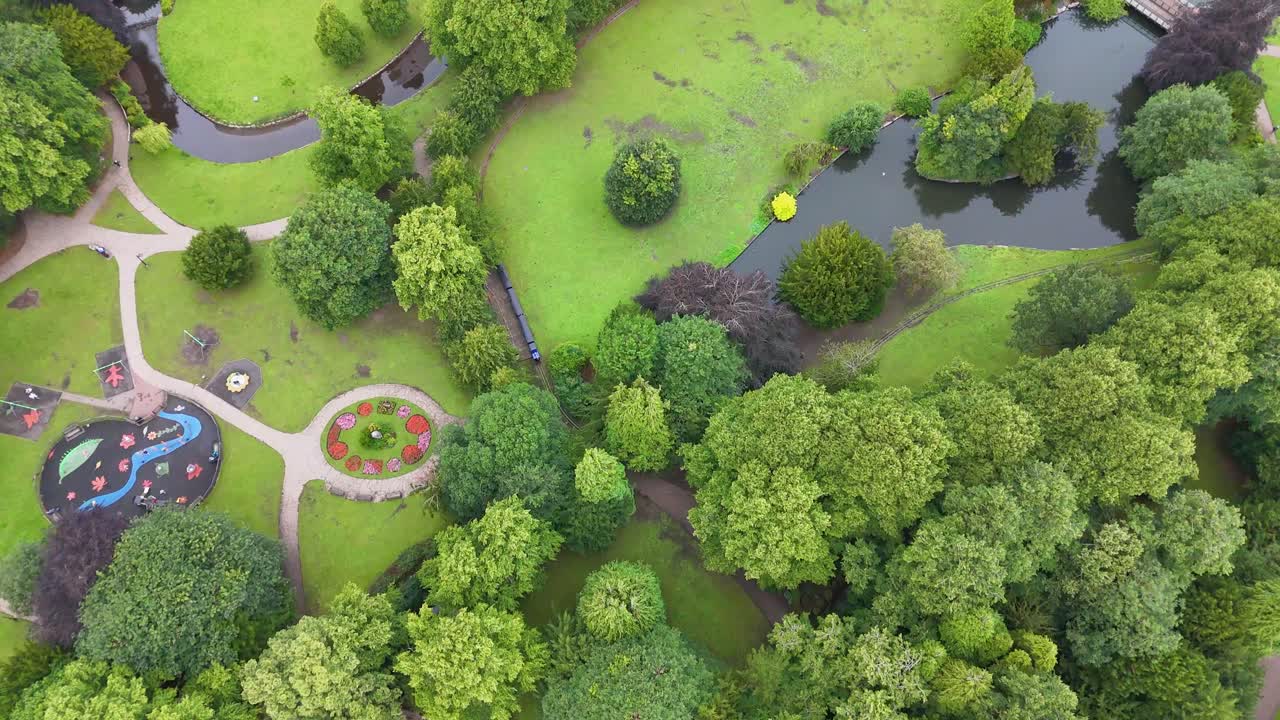 Drone camera glides above a vibrant green park with winding paths, trees, and a pond, under soft daylight, capturing a peaceful summer atmosphere