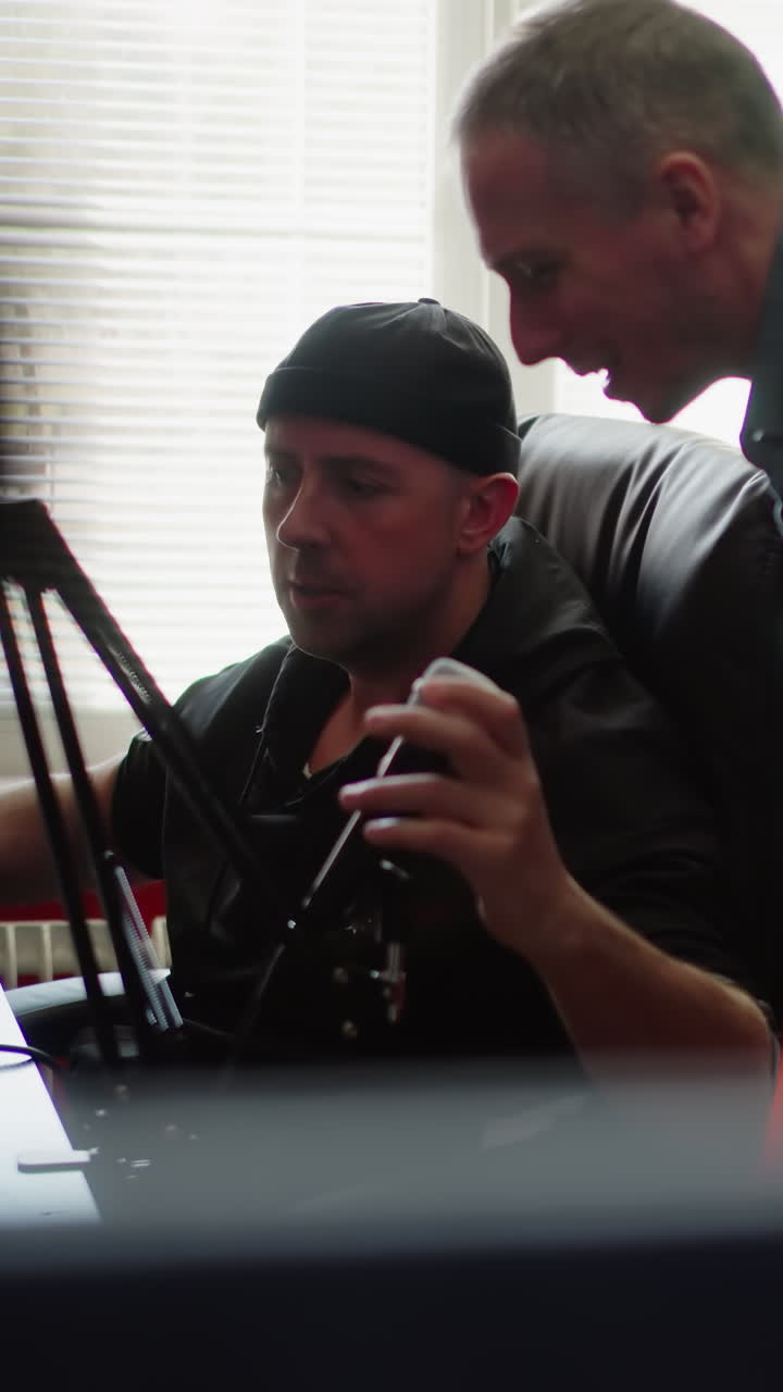 두 남자가 스튜디오 방에 앉아 있습니다. 한 명은 검은 옷을 입고 컴퓨터 앞에 앉은 프로듀서일 가능성이 높습니다. 다른 한 명은 긴 팔을 입은 셔츠를 입고 마이크를 테스트하기 위해 몸을 기울이고 있습니다.