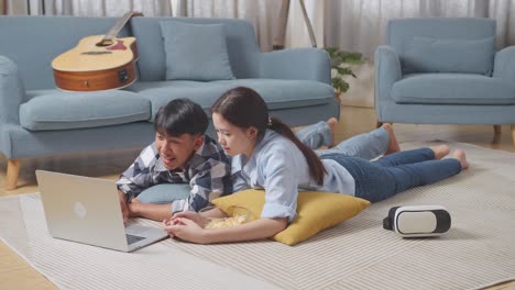 full body of asian teen couple using a laptop while lying on carpet on the floor at home. watching movie with snacks, having video call, speaking, and enjoying time together