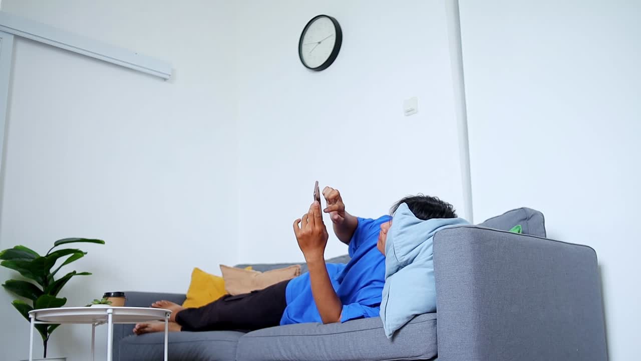 Relaxed young man scrolling smartphone on comfortable sofa enjoying modern home leisure time