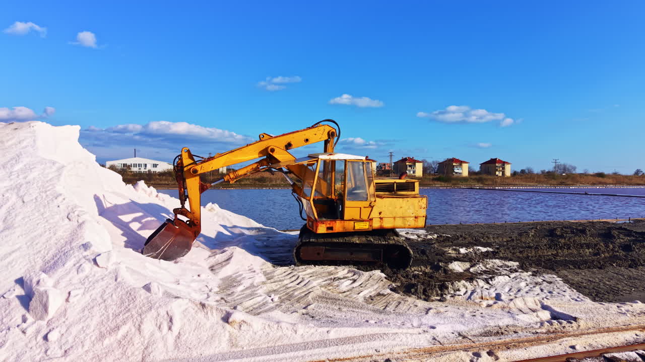 Heavy machinery moves salt along the shore under a bright blue sky with fluffy clouds, showcasing industrial activity by a tranquil water body