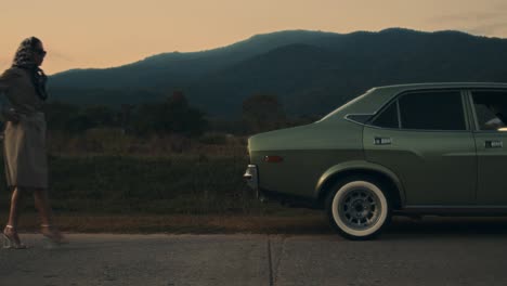 woman walking beside vintage car in rural landscape at sunset