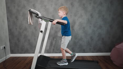 side view of little boy casually working out on treadmill reaching forward to grab cloth draped over console slow walk hold handle for balance against grey textured wall with beanbag and piano corner