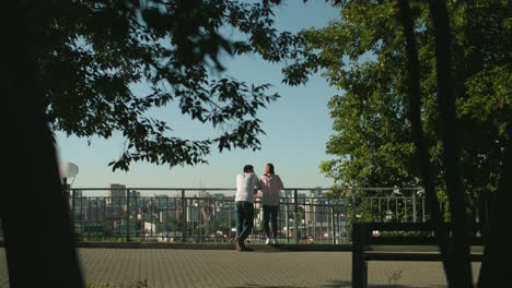 vista trasera de la pareja de pie y apoyada en una barandilla de hierro con vistas al paisaje urbano rodeado de vegetación, la mujer está de pie en el pavimento mientras el hombre la mira, bajo la cálida luz del sol