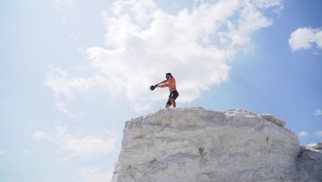 strong man lifting kettlebell on the mountain in summer. male athlete swinging kettlebell with two hands on the beautiful natural background outdoors.