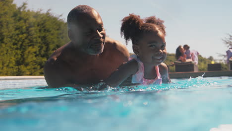 grandfather teaching granddaughter to swim in outdoor pool on holiday