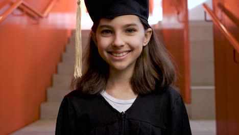 portrait of a happy preschool female student in cap and gown going down the stairs, holding graduation diploma and looking at the camera