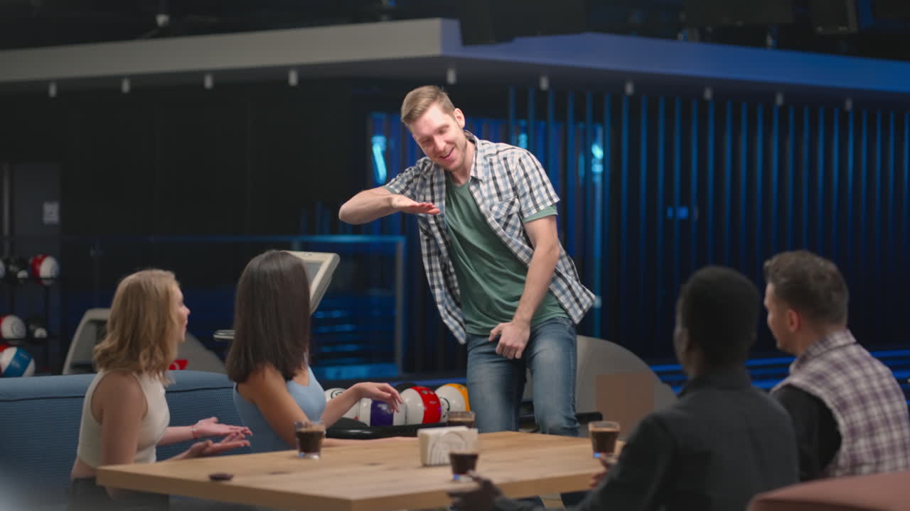 기쁜 백인 남자가 친구들에게 다섯 남녀가 볼링 클럽에서 테이블에 앉아 승리를 축하하고 팀 성공을 축하합니다.