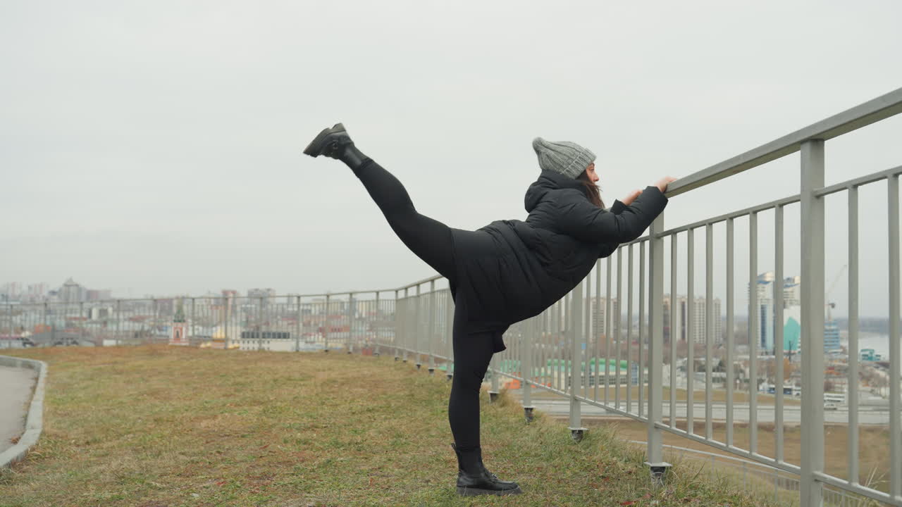 Improvvisazione di danza a bordo ferrovia con la skyline, artista solitario che prova rapidi movimenti ritmici delle gambe mentre si tiene alla ringhiera, piumino e berretto contro la brezza fredda, slancio giocoso che si trasforma in equilibrio.