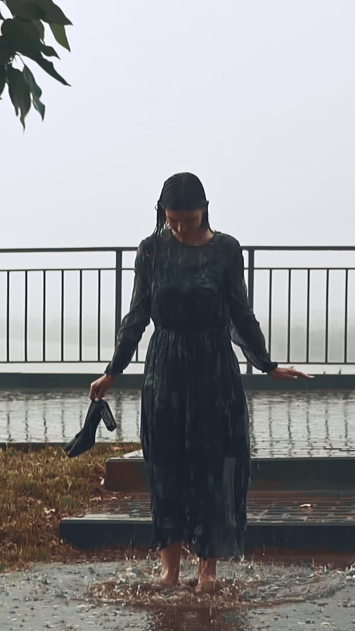 Playful young woman in wet clothes jumps holding heeled shoes into large puddle under heavy rain in city park on summer day slow motion
