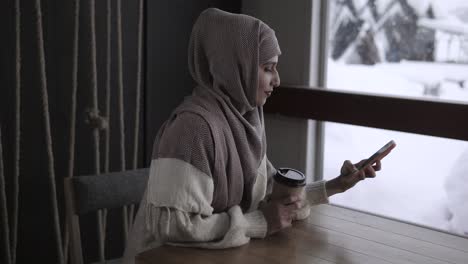 woman wearing hijab using smartphone in a cafe during winter