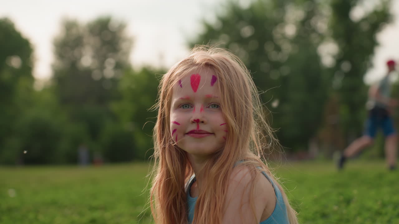 Mädchen mit Katzengesichtsbemalung sitzt lächelnd auf dem Gras, der Wind weht durch die Haare, Sommerpark-Kulisse mit sanftem Sonnenlicht, spielerischer Ausdruck, der kindliche Kreativität und Spaß im Freien auf einer grünen Wiese zeigt.