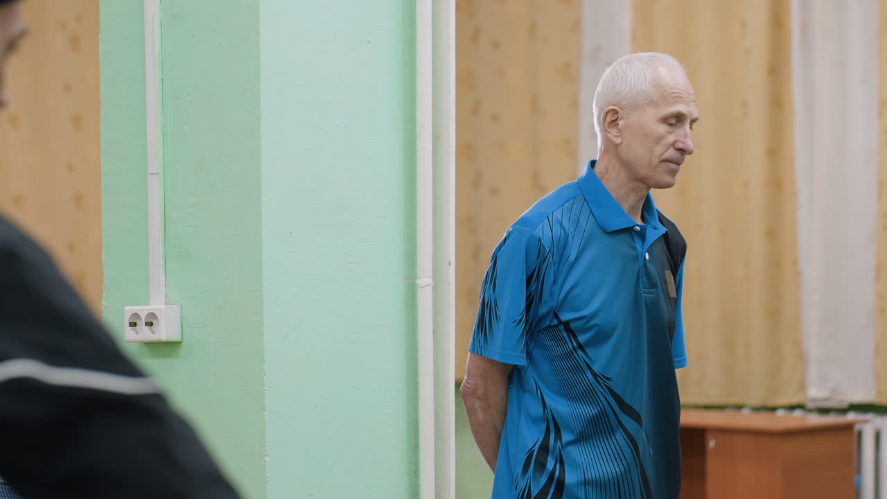 Senior trainer in blue shirt carefully watches trainee during table tennis session, focusing on performance, concentration, and skill development, emphasizing discipline, readiness, and improvement