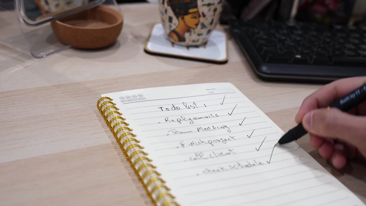 shot of a hand using a black pen to check off tasks on a handwritten to do list. The spiral notebook rests on a wooden office desk next to a keyboard and coffee mug in a modern workspace.