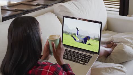 compuesto de una mujer sentada en casa sosteniendo un café viendo un juego de béisbol en una computadora portátil