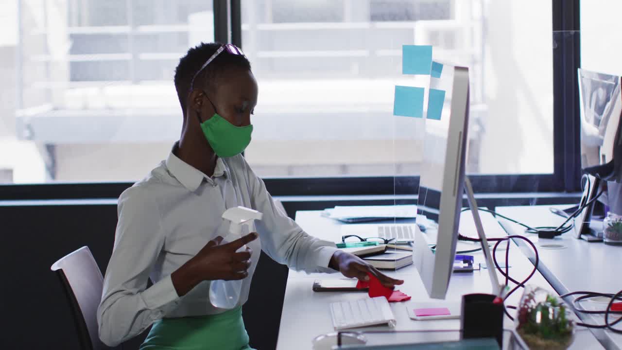 African american woman wearing face mask cleaning her desk with disinfectant at modern office