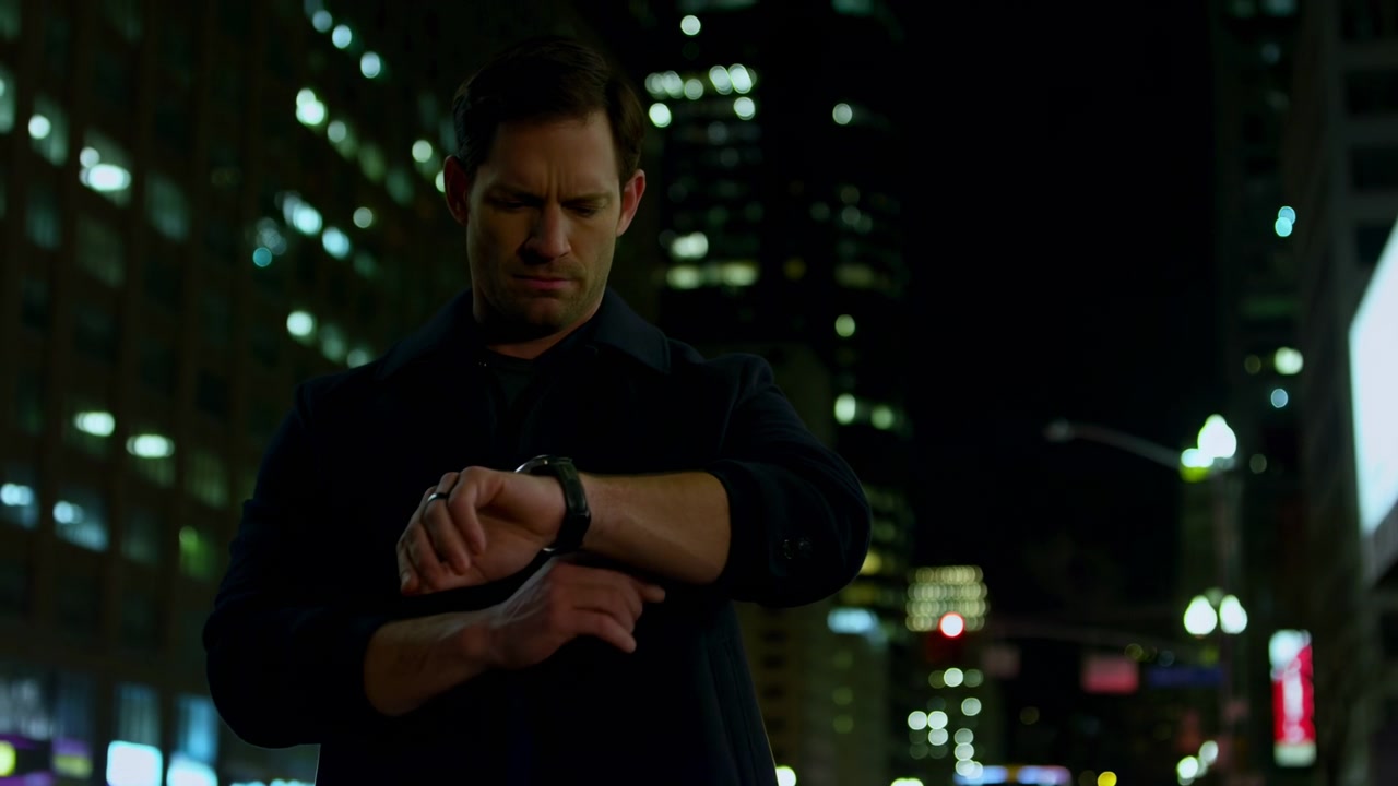 Nighttime low-angle view begins with man facing forward, then raising his wrist and looking down at his watch while soft building lights and bright panels blur behind him.