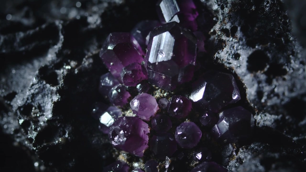 La telecamera si allontana rivelando un gruppo di cristalli viola in una configurazione macro, con la luce che si sposta per mostrare le sfaccettature.