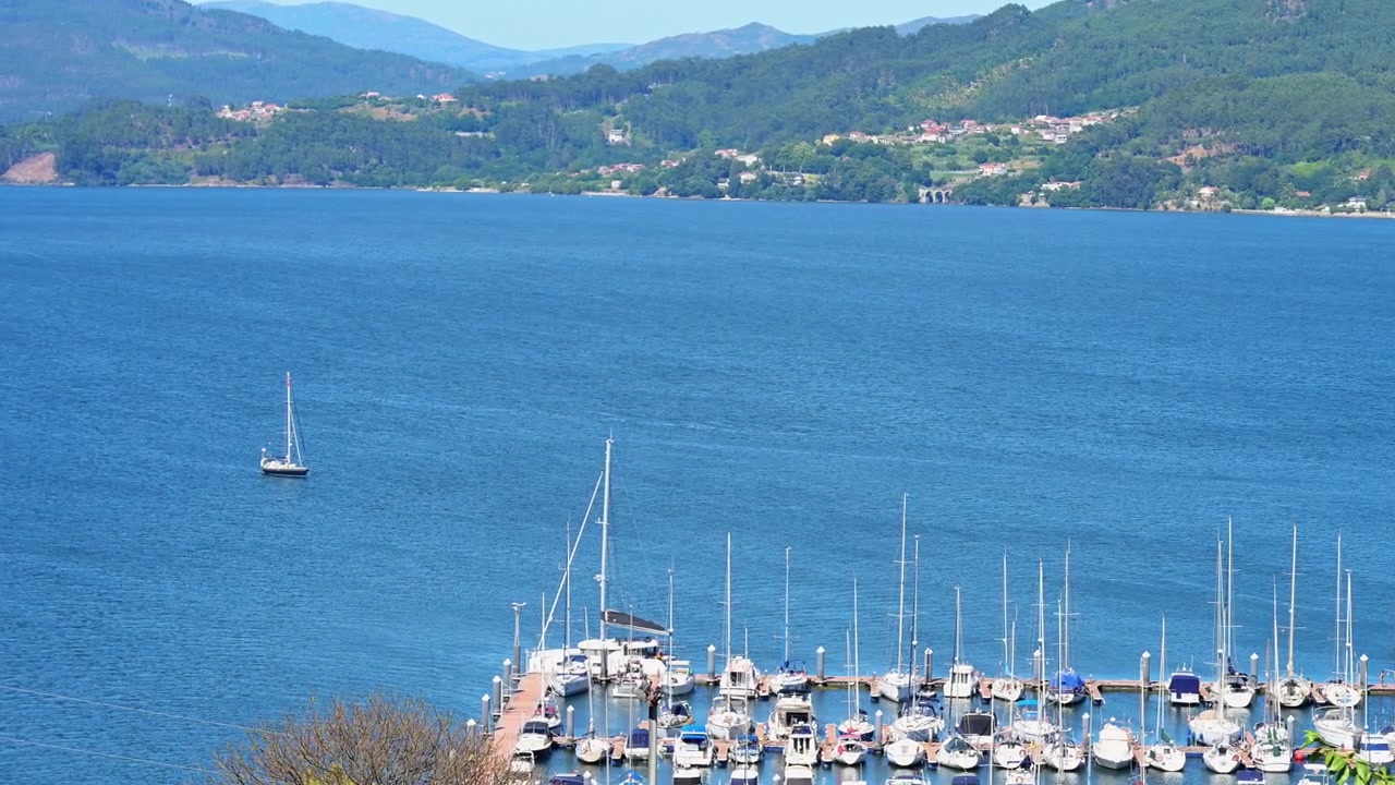 Panorama estivo soleggiato di un molo del porto turistico dell'estuario di Vigo con yacht ormeggiati e una barca a vela ancorata sull'acqua blu. Cattura il turismo nautico europeo e lo stile di vita dei resort costieri marittimi.