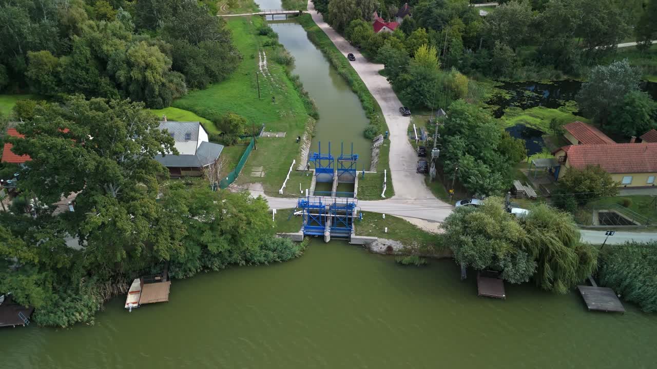 Orbital drone view of the Dömsöd sluice gate where the Danube feeds the belt canal, surrounded by riverside houses, greenery and small fishing piers