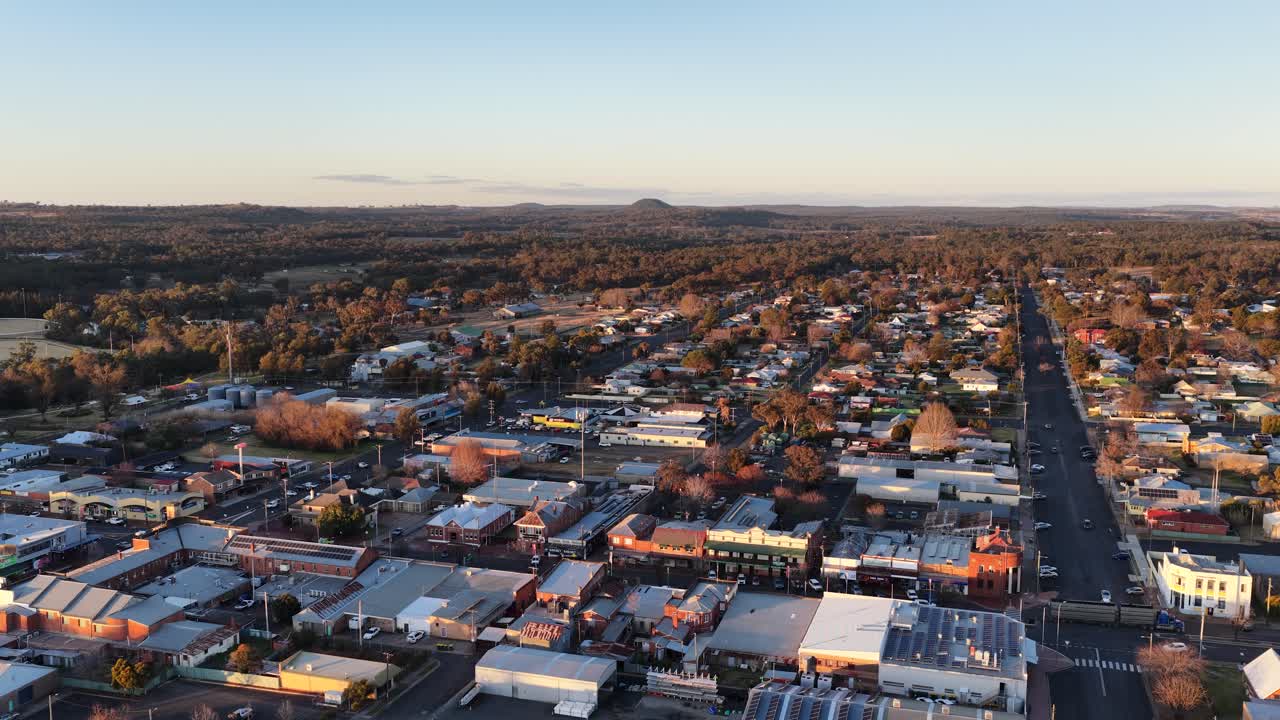 Drone glides above Coonabarabran’s main street at sunset, revealing small town buildings, tree-lined roads, and distant hills in warm, golden light