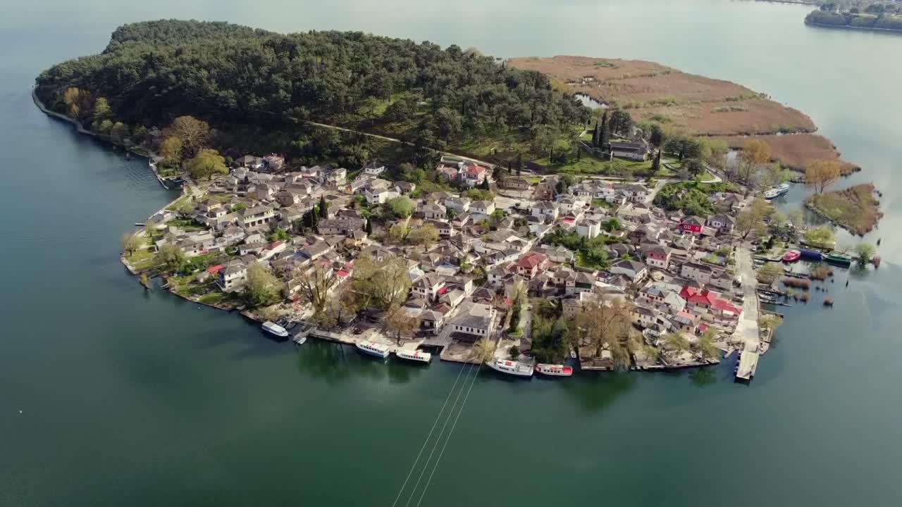 Et filmatisk luftfoto som starter fra innsjønivå og vipper opp for å avsløre steinbygningene i den tradisjonelle bosetningen på Ioannina-øya, omgitt av den rolige innsjøen Pamvotida i Hellas.