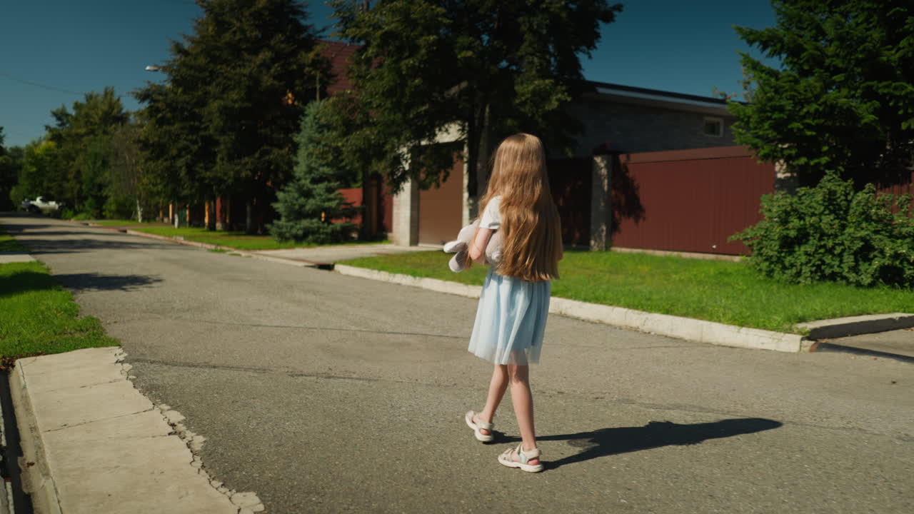 Rear view of little girl in light blue gown with net fabric walking along calm residential street as sun casts sharp shadow on pavement near green lawn and visible roadside drainage