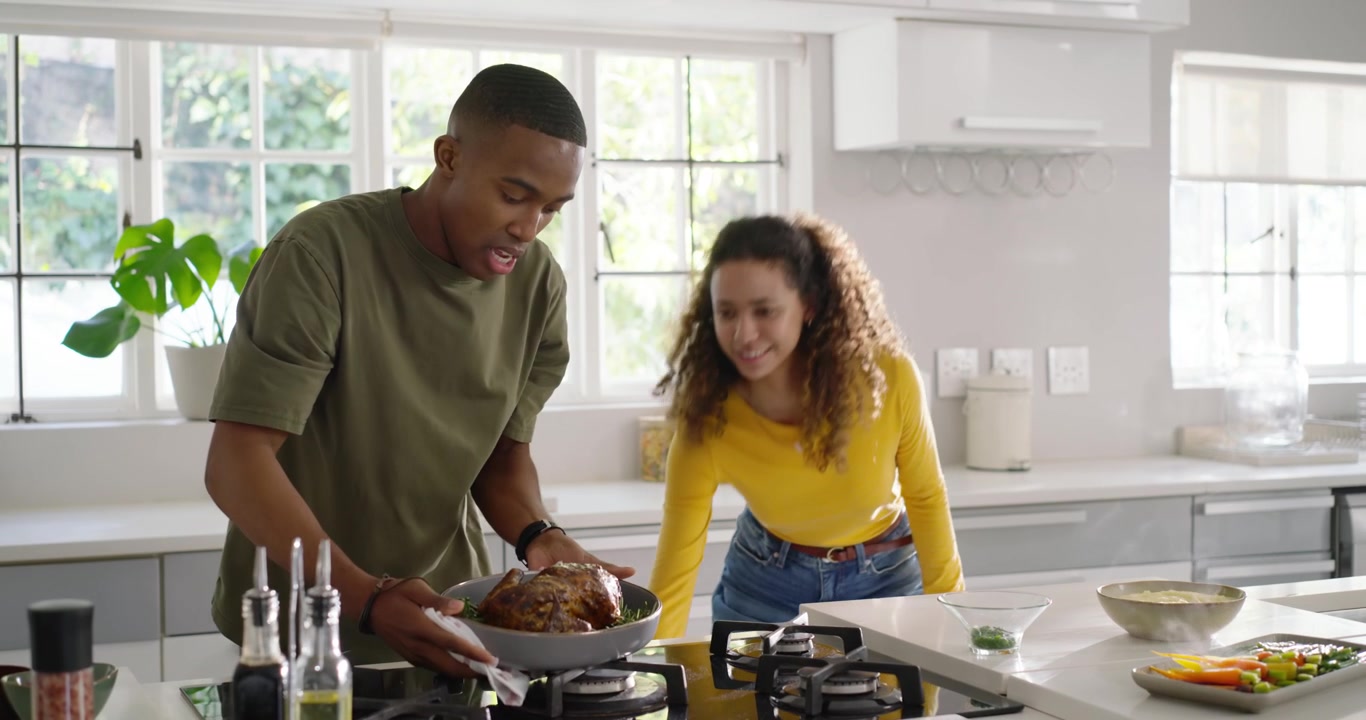 Diverse couple in home kitchen, woman entering, man holding roasting pan checking roast on gas hob
