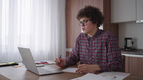 curly haired male student attractive young boy in glasses is studying at home using laptop typing writing in notebook. college student using laptop computer watching distance online learning seminar