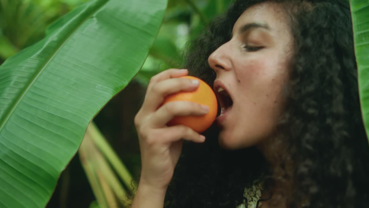 Primer plano de mujer con cabello rizado comiendo una naranja madura rodeada de hojas verdes