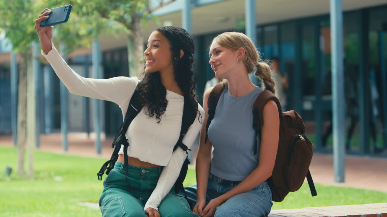 To kvinnelige videregående skoleelever som poserer for en selfie med telefonen mens de sitter på en mur utendørs
