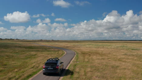 Car-driving-on-narrow-asphalt-road-winding-in-flat-countryside.-Vast-coastal-grasslands-on-sunny-day.-Denmark
