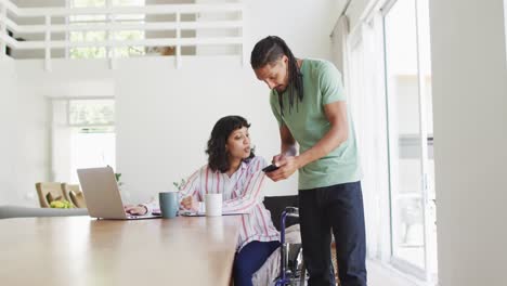 Happy-biracial-woman-in-wheelchair-using-laptop-in-living-room,-with-male-partner-using-smartphone