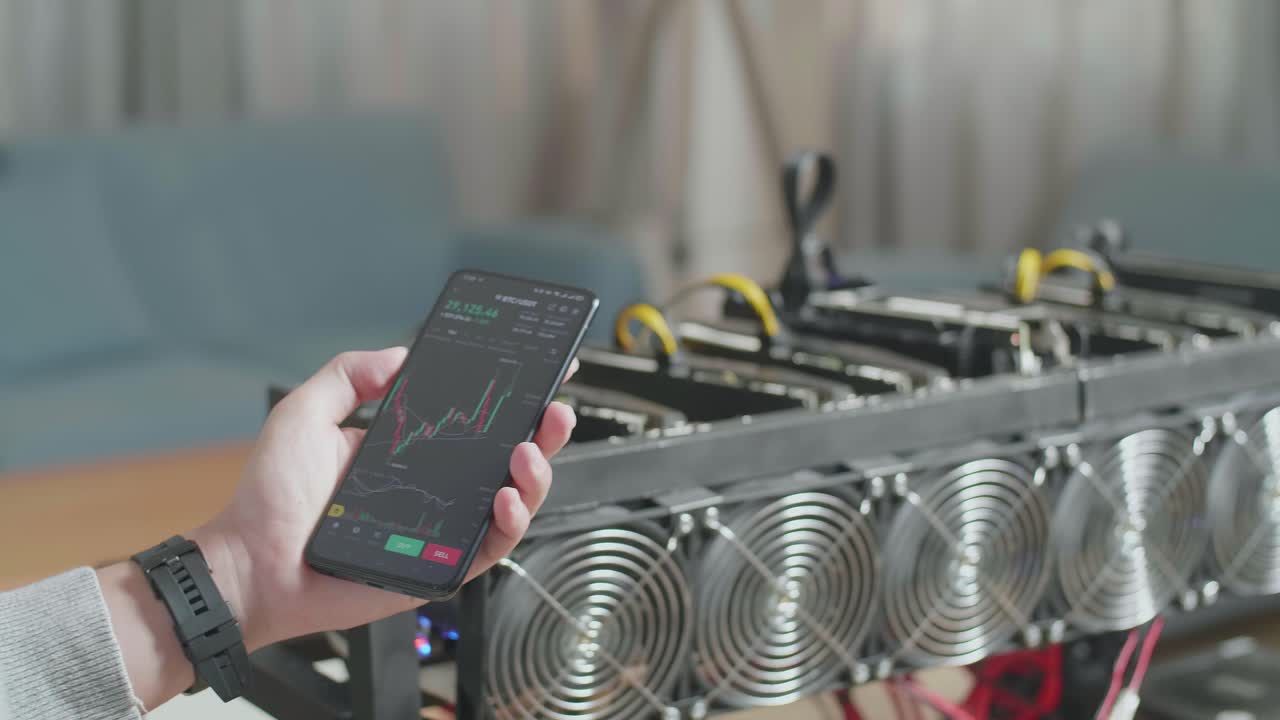 암호화폐를 채굴하기 위한 광산 기기의 앞에 비트코인 차트를 보여주는 스마트폰을 들고 있는 사람의 손의 근접 사진